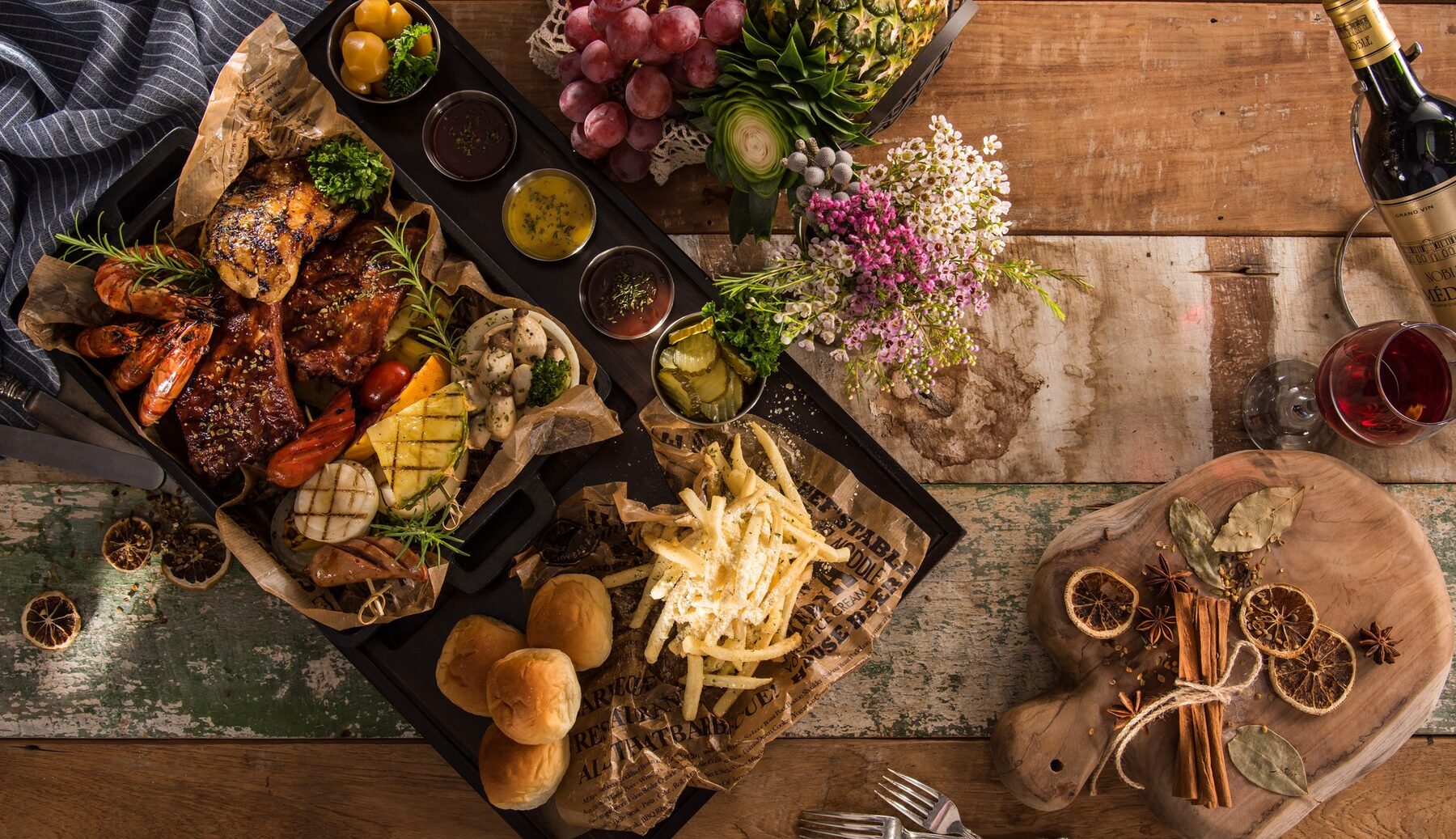 Mesa de catering con tabla de asados, frutas, flores y vino sobre madera rústica